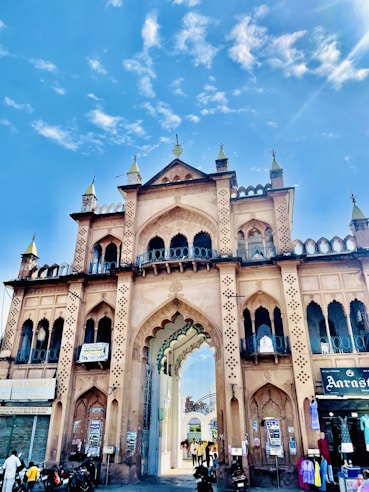 A grand historic gateway with intricate architectural details and arches stands tall against a vibrant blue sky. The structure is adorned with multiple tiers, pointed minarets, and decorative patterns. People and vehicles are seen passing through the gateway, adding a lively urban atmosphere.