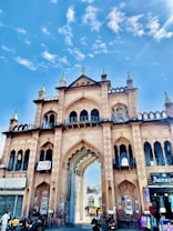 A grand historic gateway with intricate architectural details and arches stands tall against a vibrant blue sky. The structure is adorned with multiple tiers, pointed minarets, and decorative patterns. People and vehicles are seen passing through the gateway, adding a lively urban atmosphere.