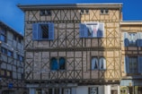 A historic building with a timber-framed facade featuring brickwork and wooden beams. The structure has multiple blue window shutters, some open and some closed. Below, there is a hair salon with signage reading 'Jean Louis David'. The building is set against a clear blue sky.