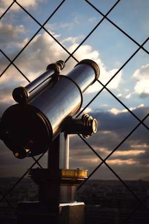 A metallic telescope mounted on a stand is facing towards a sky filled with clouds and a setting sun, framed by a metal grid. The telescope has shiny brass and silver elements, reflecting the soft light.