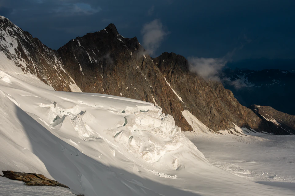 The image depicts a majestic mountain range with snow-capped peaks and a deep blue sky. The mountains, painted in shades of brown and gray, rise high into the sky, their rocky surfaces contrasting against the white snow that blankets them. The perspective is from below, looking up at these towering natural wonders. In the foreground, there's a small patch of grass, adding a touch of green to the otherwise monochrome landscape.