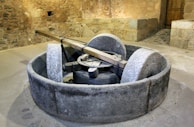 An old millstone setup situated in a rustic stone-walled room. The grinding apparatus consists of two large stone wheels inside a circular stone basin. A wooden beam extends above the stones, indicating manual or animal-powered operation. The walls and floor are made from natural stone, enhancing the historical ambiance.