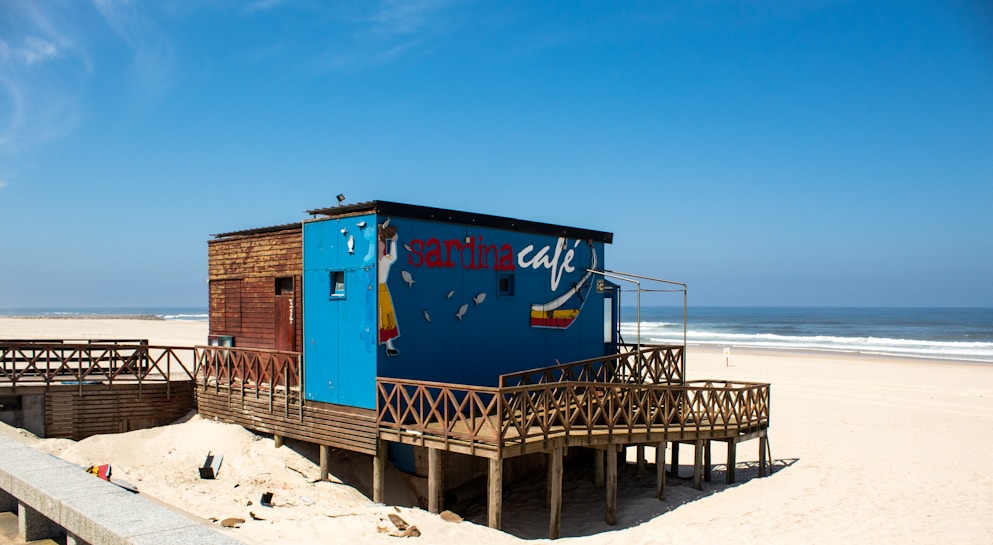 A quaint beachside cafe with a wooden exterior and a vibrant blue section featuring artistic designs and the words 'sardina cafe'. It is elevated on stilts over the sandy beach, with a wooden railing surrounding the structure. The ocean with gentle waves and a clear sky provides a serene backdrop.