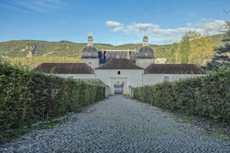 A long cobblestone path leads to a historic building with large, distinctive domes set against a backdrop of lush green mountains and a clear blue sky. Dense hedges line either side of the path, and the entrance to the building features an arched gateway.