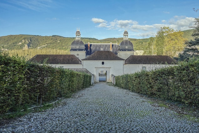 A long cobblestone path leads to a historic building with large, distinctive domes set against a backdrop of lush green mountains and a clear blue sky. Dense hedges line either side of the path, and the entrance to the building features an arched gateway.