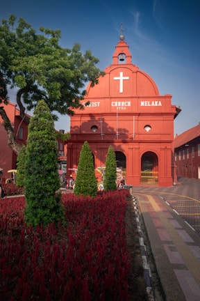 A vibrant red historical church building with a prominent white cross at the top. The church, named Christ Church, is dated 1753 and features a Dutch colonial architectural style. In front of the building, there are meticulously arranged red flowers and tall green shrubs that add to the aesthetic appeal. A few colorful trishaws can be seen near the entrance, adding cultural context.