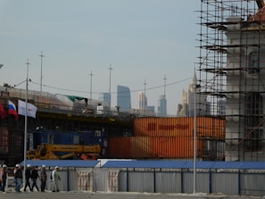 A construction site is visible with scaffolding and construction materials. Several workers wearing hard hats are walking along a pathway. Flags are displayed, and tall modern buildings can be seen in the distant skyline.