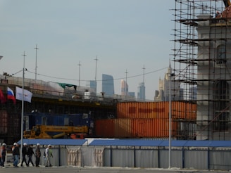 A construction site is visible with scaffolding and construction materials. Several workers wearing hard hats are walking along a pathway. Flags are displayed, and tall modern buildings can be seen in the distant skyline.