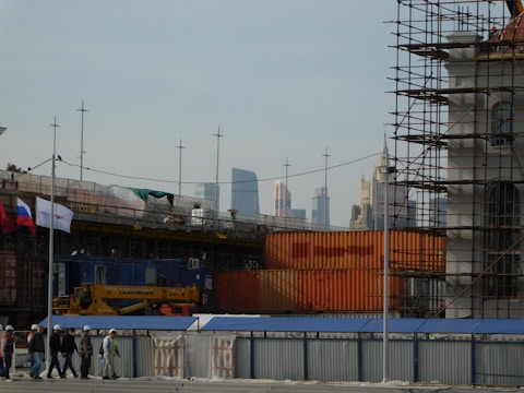 A construction site is visible with scaffolding and construction materials. Several workers wearing hard hats are walking along a pathway. Flags are displayed, and tall modern buildings can be seen in the distant skyline.