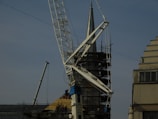 A construction site featuring a large crane with a visible metal arm dominating the scene. Scaffolding surrounds a pointed tower or spire, suggesting some type of renovation work. Workers are at the base, wearing safety gear, and several ropes and cables are seen hanging from the crane.