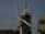 A construction site featuring a large crane with a visible metal arm dominating the scene. Scaffolding surrounds a pointed tower or spire, suggesting some type of renovation work. Workers are at the base, wearing safety gear, and several ropes and cables are seen hanging from the crane.