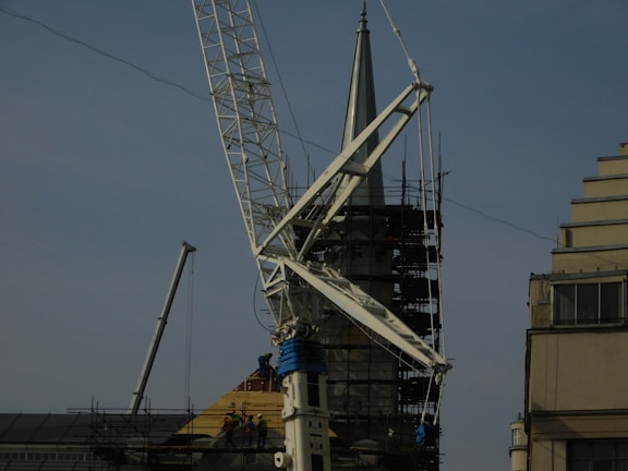 A construction site featuring a large crane with a visible metal arm dominating the scene. Scaffolding surrounds a pointed tower or spire, suggesting some type of renovation work. Workers are at the base, wearing safety gear, and several ropes and cables are seen hanging from the crane.
