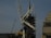 A construction site featuring a large crane with a visible metal arm dominating the scene. Scaffolding surrounds a pointed tower or spire, suggesting some type of renovation work. Workers are at the base, wearing safety gear, and several ropes and cables are seen hanging from the crane.