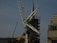 A construction site featuring a large crane with a visible metal arm dominating the scene. Scaffolding surrounds a pointed tower or spire, suggesting some type of renovation work. Workers are at the base, wearing safety gear, and several ropes and cables are seen hanging from the crane.