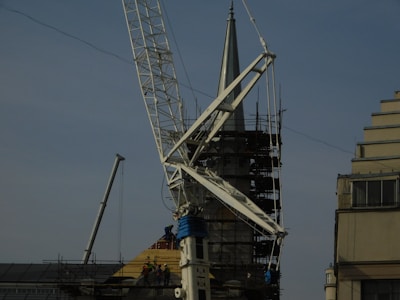 A construction site featuring a large crane with a visible metal arm dominating the scene. Scaffolding surrounds a pointed tower or spire, suggesting some type of renovation work. Workers are at the base, wearing safety gear, and several ropes and cables are seen hanging from the crane.
