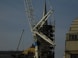 A construction site featuring a large crane with a visible metal arm dominating the scene. Scaffolding surrounds a pointed tower or spire, suggesting some type of renovation work. Workers are at the base, wearing safety gear, and several ropes and cables are seen hanging from the crane.