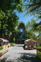 A modern building with a large glass facade is surrounded by lush green trees. In front of the building, tents are set up along the walkway, with people engaging in activities under them. The area is well-lit by sunlight, enhancing the vibrant atmosphere.