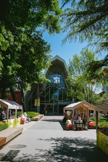 A modern building with a large glass facade is surrounded by lush green trees. In front of the building, tents are set up along the walkway, with people engaging in activities under them. The area is well-lit by sunlight, enhancing the vibrant atmosphere.