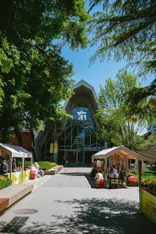 A modern building with a large glass facade is surrounded by lush green trees. In front of the building, tents are set up along the walkway, with people engaging in activities under them. The area is well-lit by sunlight, enhancing the vibrant atmosphere.