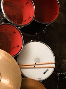 A set of drums with red drum heads and a white snare drum is prominently visible. Two drumsticks are resting on the snare drum. A golden cymbal is also partially visible in the foreground. The image gives a close-up view from an overhead angle.