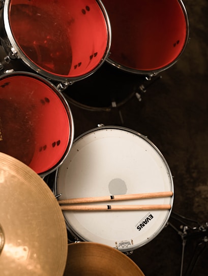 A set of drums with red drum heads and a white snare drum is prominently visible. Two drumsticks are resting on the snare drum. A golden cymbal is also partially visible in the foreground. The image gives a close-up view from an overhead angle.