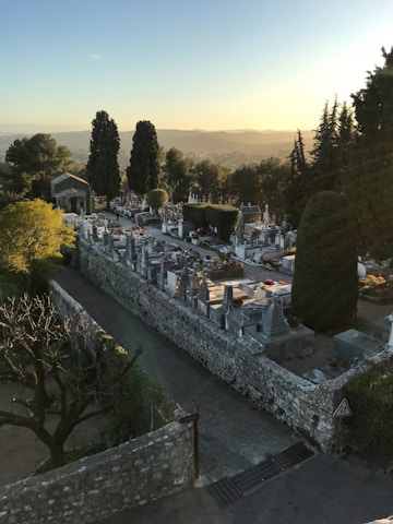 A serene cemetery surrounded by lush greenery and tall trees under a clear blue sky during sunset. Well-maintained graves and tombstones line a stone path, with a small chapel visible in the distance. The scene is tranquil, with soft, warm lighting from the setting sun casting long shadows.