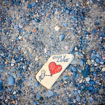 A painted rock rests among a collection of small pebbles. The rock displays a drawing of a person holding a red heart-shaped balloon with the words 'ONLY LOVE' written above.