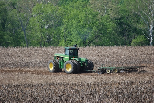 A green tractor is working in a brown, plowed field, surrounded by lush green trees in the background. The tractor appears to be tilling or cultivating the soil, with a piece of agricultural machinery attached at the back, stirring up the earth as it moves.