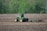 A green tractor is working in a brown, plowed field, surrounded by lush green trees in the background. The tractor appears to be tilling or cultivating the soil, with a piece of agricultural machinery attached at the back, stirring up the earth as it moves.