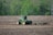 A green tractor is working in a brown, plowed field, surrounded by lush green trees in the background. The tractor appears to be tilling or cultivating the soil, with a piece of agricultural machinery attached at the back, stirring up the earth as it moves.