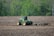A green tractor is working in a brown, plowed field, surrounded by lush green trees in the background. The tractor appears to be tilling or cultivating the soil, with a piece of agricultural machinery attached at the back, stirring up the earth as it moves.