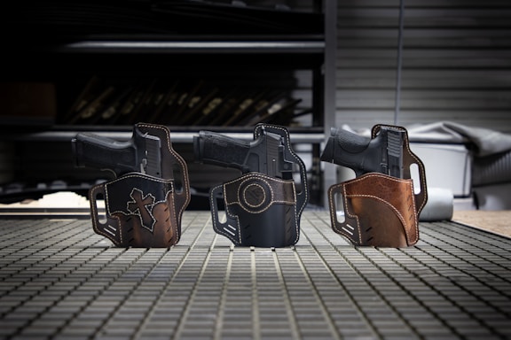 Three handguns are securely placed in leather holsters on a grid-patterned surface. Each holster is uniquely designed with stitching details and distinct patches, including a cross on the left holster. The background includes shelves with various items, out of focus, in a workshop-like setting.