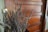 A bundle of dried lavender is arranged in front of a wooden paneled door. The lavender appears slightly wilted with muted purple hues, creating a rustic and calming atmosphere. The wooden door has a rich, warm brown color with visible grains and a brass handle.