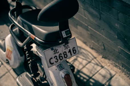 A close-up view of a parked motorbike with a license plate reading 'C36816', situated against a brick wall. The motorbike is largely black and white, featuring a sturdy seat and a handle or guard with the word 'STRICT'. Sunlight casts shadows onto the pavement surrounding the bike.