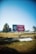 A large billboard stands in an open, grassy area under a clear blue sky. The billboard features a Coca-Cola advertisement with the text 'Wishing You a Safe Journey' and some imagery. Trees and a parked car are visible in the background, adding to the spacious outdoor setting.