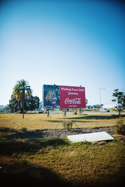 A large billboard stands in an open, grassy area under a clear blue sky. The billboard features a Coca-Cola advertisement with the text 'Wishing You a Safe Journey' and some imagery. Trees and a parked car are visible in the background, adding to the spacious outdoor setting.