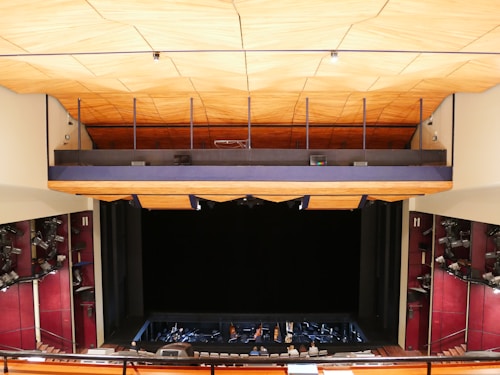 An auditorium or theater interior with a large stage at the bottom. The space is elevated above the stage, likely for seats or equipment. The wood-paneled ceiling creates a warm atmosphere, and the stage lights on the sides are directed towards the stage area.