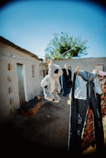 A clothesline is strung across an outdoor area, hanging worn, ripped jeans, a stuffed animal, colorful floral fabric, and a pair of socks. The background features a rustic building with a corrugated metal roof, and a tree with green leaves under a bright blue sky.