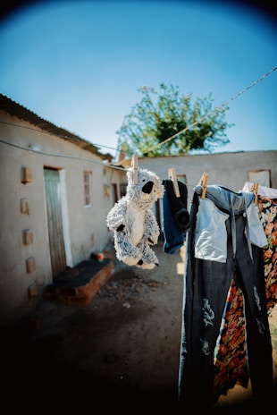 A clothesline is strung across an outdoor area, hanging worn, ripped jeans, a stuffed animal, colorful floral fabric, and a pair of socks. The background features a rustic building with a corrugated metal roof, and a tree with green leaves under a bright blue sky.