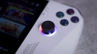 A close-up view of a handheld gaming device focusing on the joystick and buttons. The joystick is illuminated with a gradient of colors, and four colored buttons are labeled with letters. Part of a screen displaying a game is partially visible.