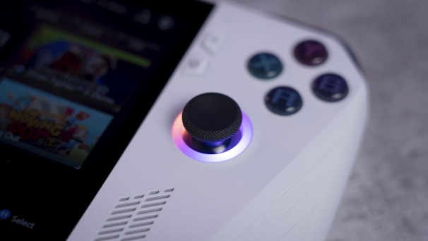 A close-up view of a handheld gaming device focusing on the joystick and buttons. The joystick is illuminated with a gradient of colors, and four colored buttons are labeled with letters. Part of a screen displaying a game is partially visible.