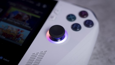 A close-up view of a handheld gaming device focusing on the joystick and buttons. The joystick is illuminated with a gradient of colors, and four colored buttons are labeled with letters. Part of a screen displaying a game is partially visible.