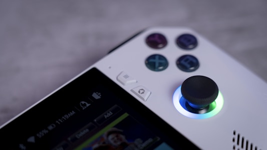 A close-up view of a gaming console controller featuring buttons with directional arrows and symbols. A prominent joystick is illuminated with a blue light. The edges of a screen display various icons and a time of 11:19AM with a battery indicator showing 55% charge.