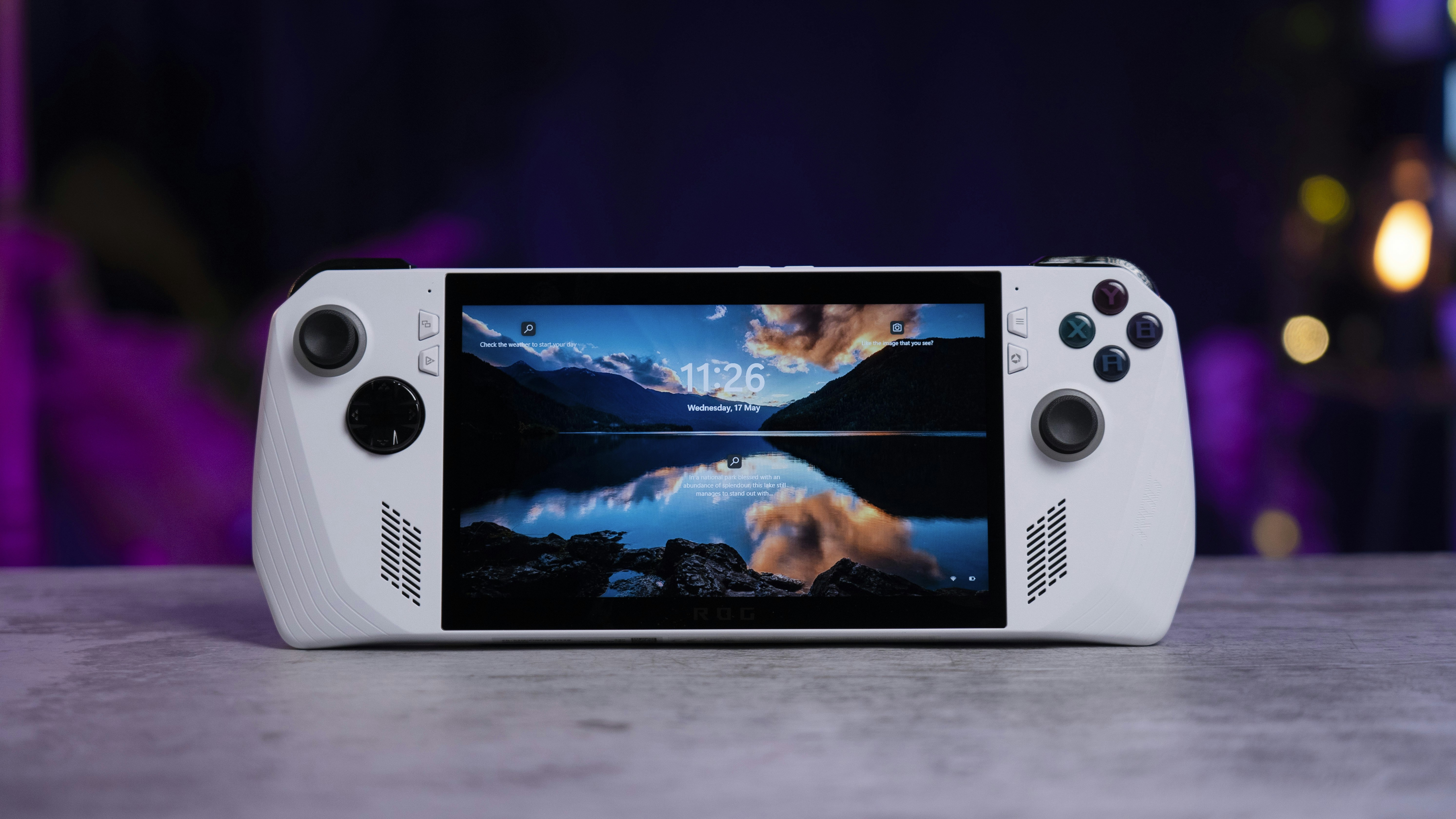 A handheld gaming console with a central screen displaying a scenic wallpaper of mountains and a lake, flanked by controller buttons and joysticks on both sides. The lighting around is dim, with purplish hues in the background, creating a modern and elegant atmosphere.
