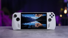 A handheld gaming console with a central screen displaying a scenic wallpaper of mountains and a lake, flanked by controller buttons and joysticks on both sides. The lighting around is dim, with purplish hues in the background, creating a modern and elegant atmosphere.