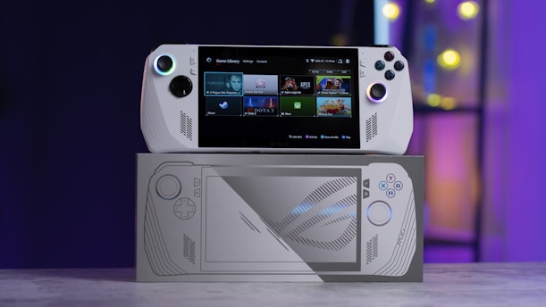 A handheld gaming console with a screen displaying a game library menu is placed on top of its packaging. The console features various buttons, a joystick, and a circular light around the left joystick. The background is blurred with purple, yellow, and blue tones.