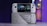 A handheld gaming console with a screen displaying a game library menu is placed on top of its packaging. The console features various buttons, a joystick, and a circular light around the left joystick. The background is blurred with purple, yellow, and blue tones.
