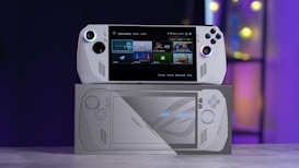 A handheld gaming console with a screen displaying a game library menu is placed on top of its packaging. The console features various buttons, a joystick, and a circular light around the left joystick. The background is blurred with purple, yellow, and blue tones.