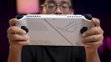 A person holds a white handheld gaming device with black controls, featuring a futuristic design on its back. The person is focused on the device with a blurred background creating a sense of depth.