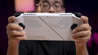 A person holds a white handheld gaming device with black controls, featuring a futuristic design on its back. The person is focused on the device with a blurred background creating a sense of depth.